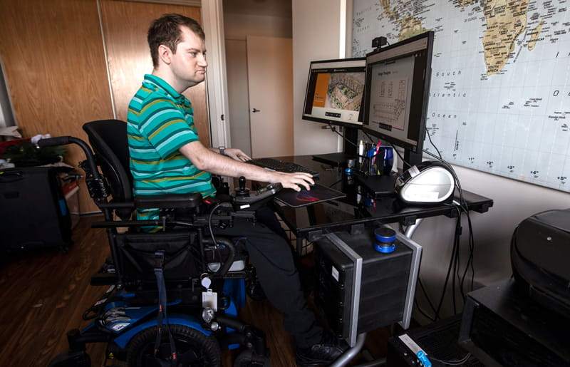 Levi Lawton seated in powerchair looks at housing designs on his computer screen. A world map is on the wall behind the screens.